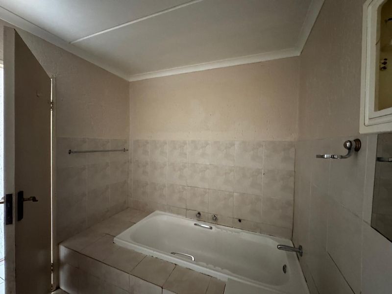 A dated bathroom featuring a built-in bathtub with tiled surround, wall-mounted fixtures, and neutral wall finishes, requiring cosmetic updates to modernize the space.