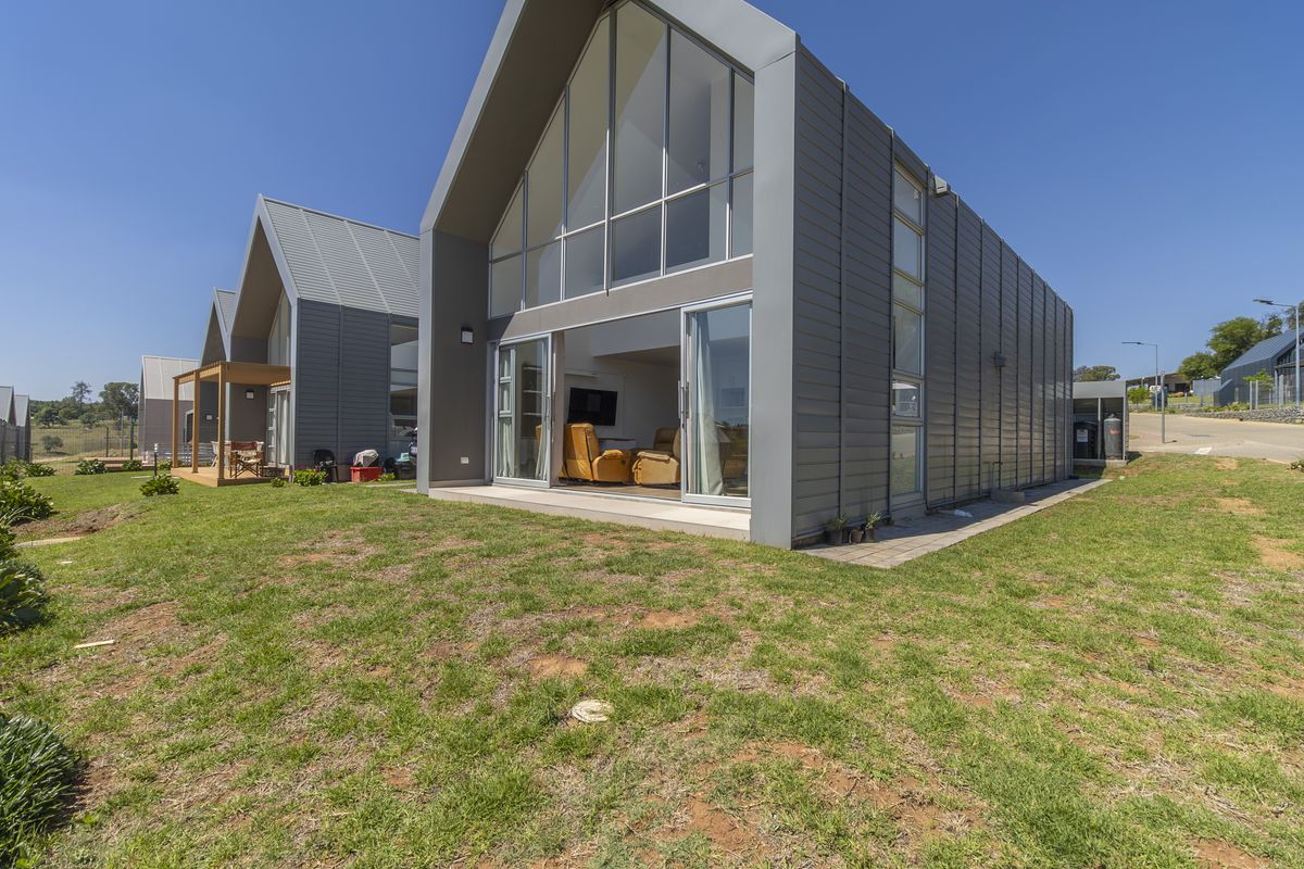Modern architectural rear elevation featuring striking metal cladding, expansive floor-to-ceiling glass gable windows, and a seamless transition to a paved patio area.