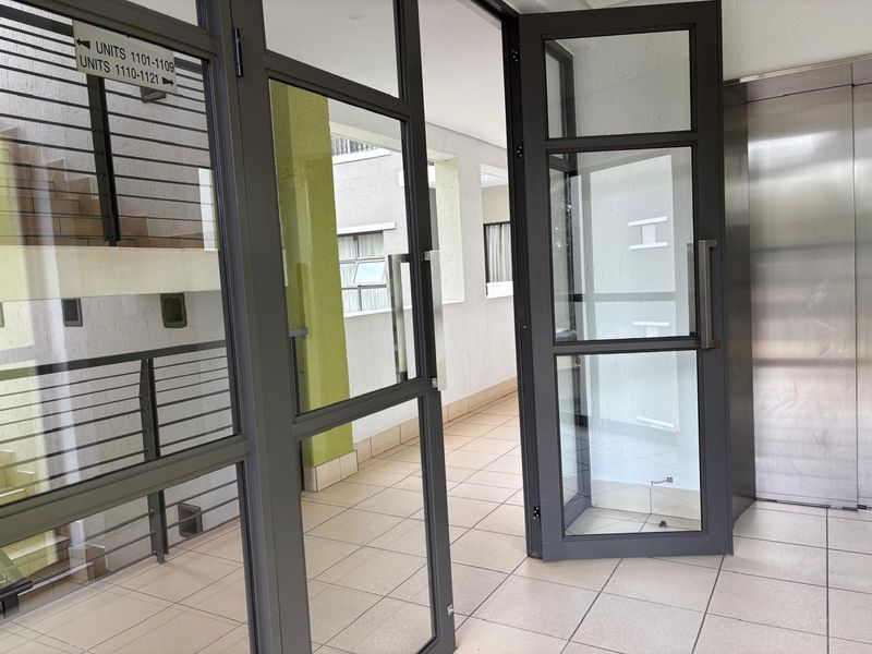 A clean, modern apartment building hallway featuring tiled floors, aluminium glass doors, and access to an elevator and stairwell.