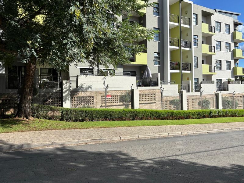 Exterior view of a modern apartment complex featuring balconies, secure brick boundary walling with electric fencing, and a well-maintained sidewalk.