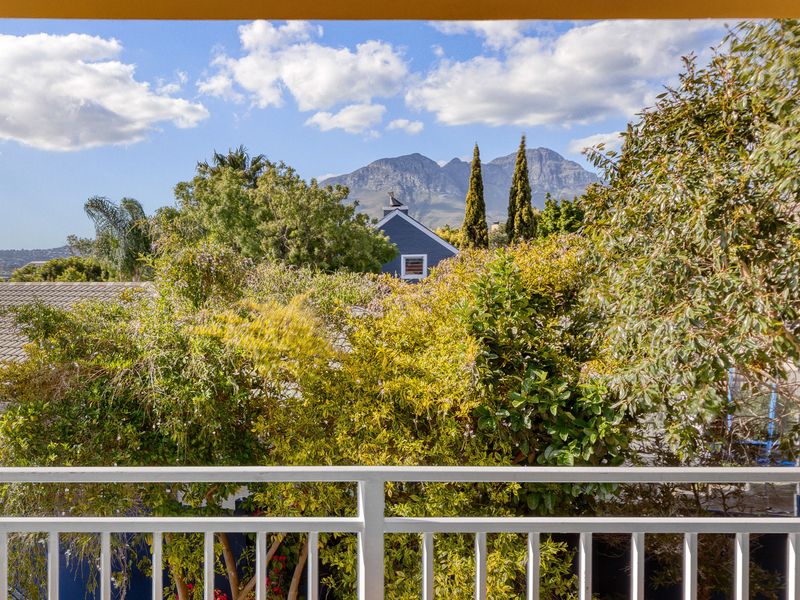 A scenic elevated view overlooking lush garden greenery and neighbouring rooftops, framed by a white metal balcony railing with mountain vistas in the background.