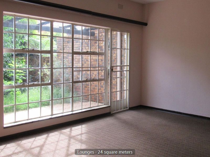 Spacious living area featuring large steel-framed windows, a security gate, and carpeted flooring, offering direct access to the outdoor patio area.