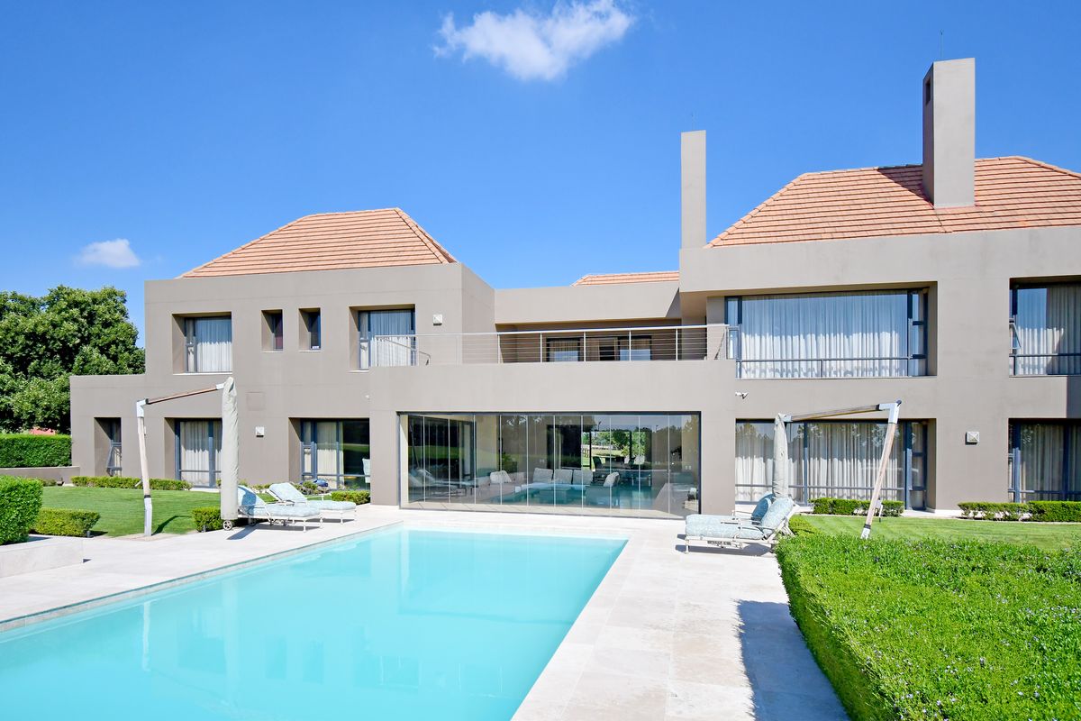 Rear view of a modern luxury residence featuring a swimming pool, stone patio, landscaped garden, and an upper-level balcony with glass sliding doors.