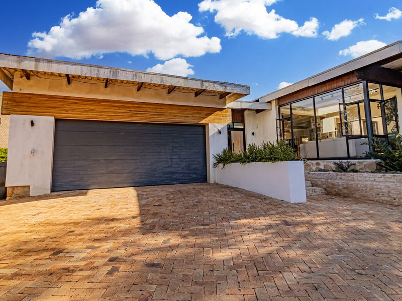 Front elevation featuring a brick paved driveway, modern sectional garage door, stone cladding accents, and large steel-framed windows.