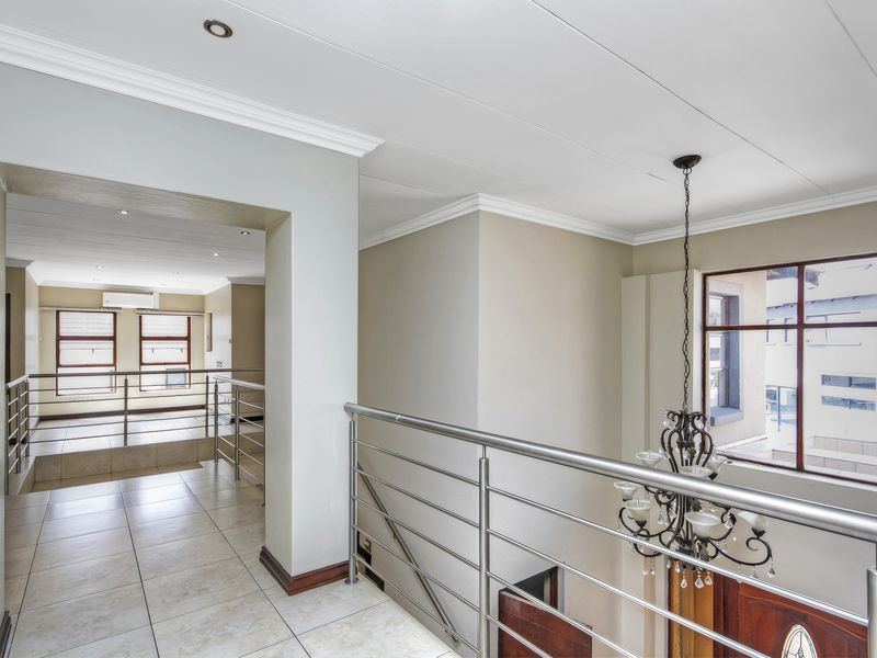 A bright and spacious upper-level landing featuring tiled flooring, a modern stainless steel balustrade, and a decorative chandelier overlooking the entrance area.