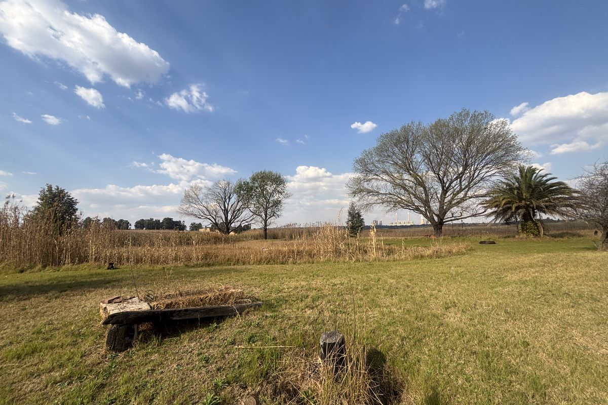 A wide-angle view of a vacant residential plot featuring open grassland, mature trees, and natural landscaping under a clear blue sky.