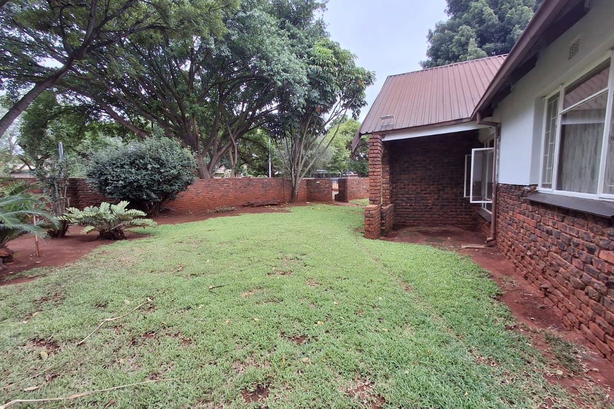 A rear garden view featuring a lawn area, mature trees, and a house exterior with exposed brick walls and a metal roof.
