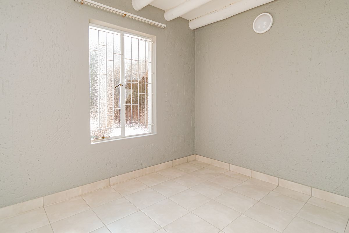 A vacant bedroom featuring neutral tiled flooring, textured walls, exposed ceiling beams, and a window fitted with security bars.