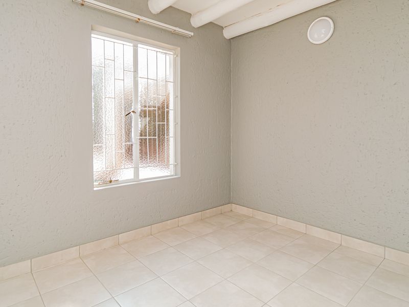 A vacant bedroom featuring neutral tiled flooring, textured walls, exposed ceiling beams, and a window fitted with security bars.