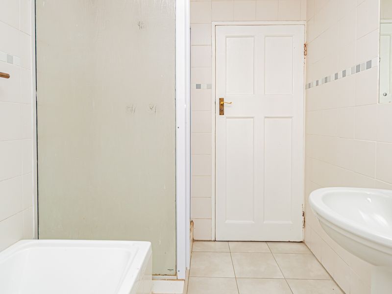A dated bathroom featuring a pedestal basin, shower enclosure, and neutral wall tiling, presenting an opportunity for modernization and cosmetic updates.