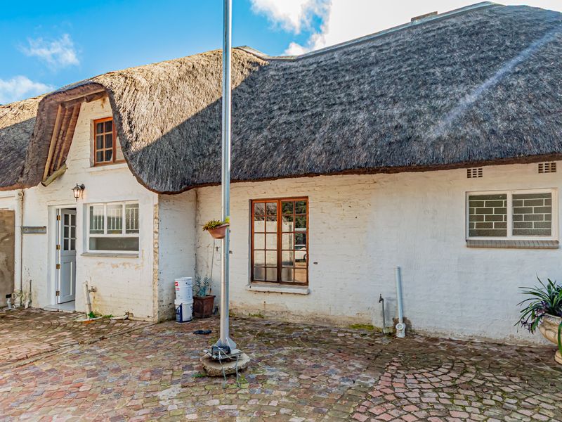Rear view of a traditional cottage featuring a thatched roof, white painted brick walls, and a brick-paved courtyard area.