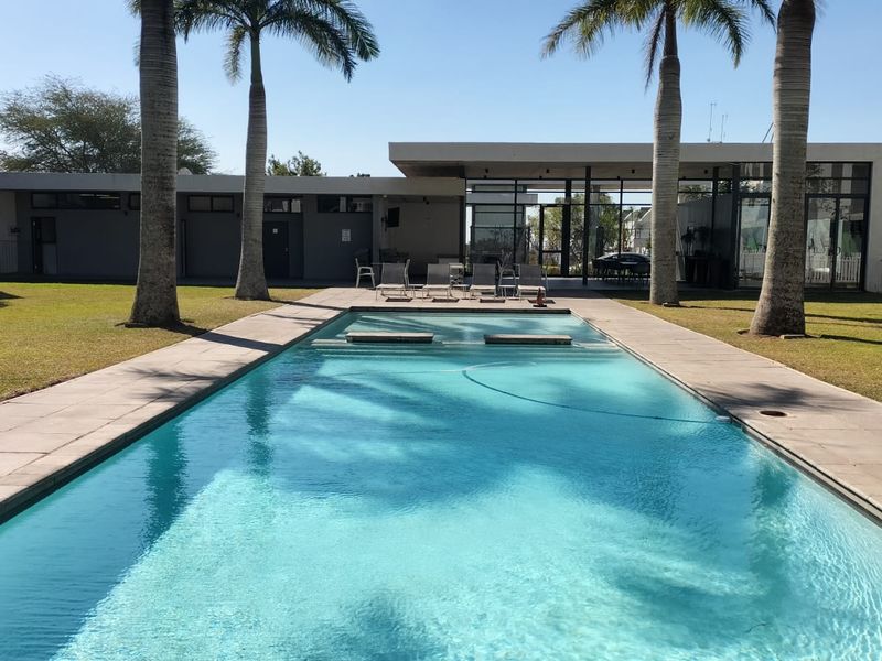 A well-maintained communal swimming pool area featuring paved decking, landscaped lawns, and palm trees, set within a modern apartment complex.