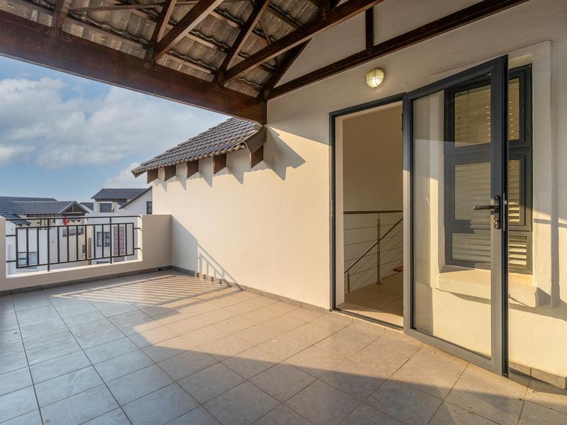 A spacious covered balcony featuring tiled flooring, exposed timber roof trusses, and a modern aluminium glass door leading into the apartment.