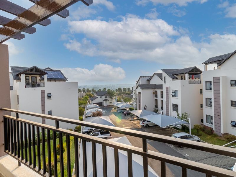 A balcony featuring a metal balustrade and timber pergola, offering an elevated view over the apartment complex grounds and parking area.