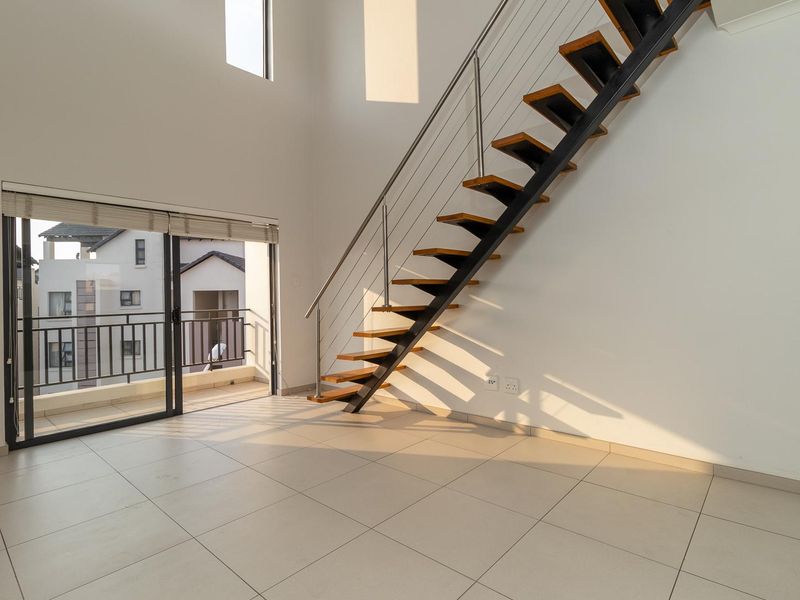 Bright, open-plan living area featuring a modern floating staircase with stainless steel balustrades, tiled flooring, and direct access to a private balcony.