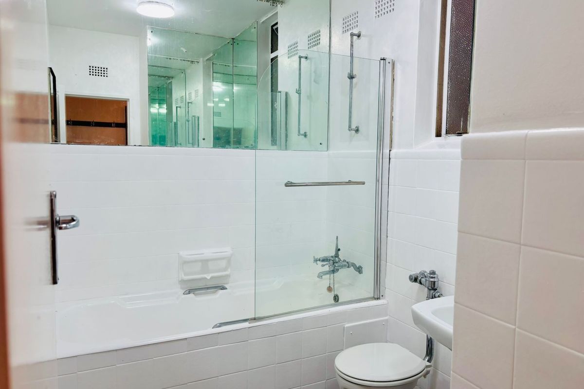 A functional bathroom featuring a white bathtub with a glass shower screen, wall-mounted basin, and toilet, finished with white wall tiles.