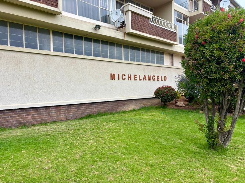 Exterior view of the Michelangelo apartment complex, featuring a landscaped lawn, brick and plaster facade, and balcony access.