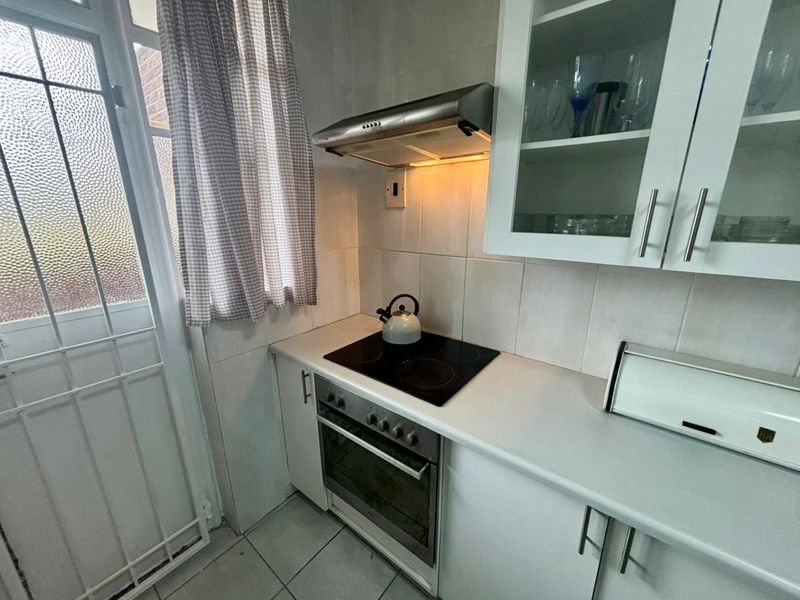 Compact kitchen featuring white cabinetry, a built-in electric oven, glass cooktop, stainless steel extractor fan, and tiled flooring with direct access to an exterior door.