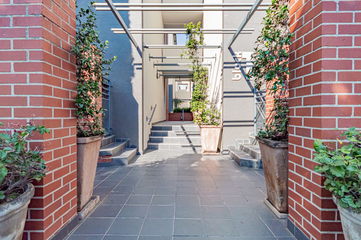 A well-maintained apartment complex entrance featuring brick pillars, a tiled walkway, and a modern metal pergola structure.