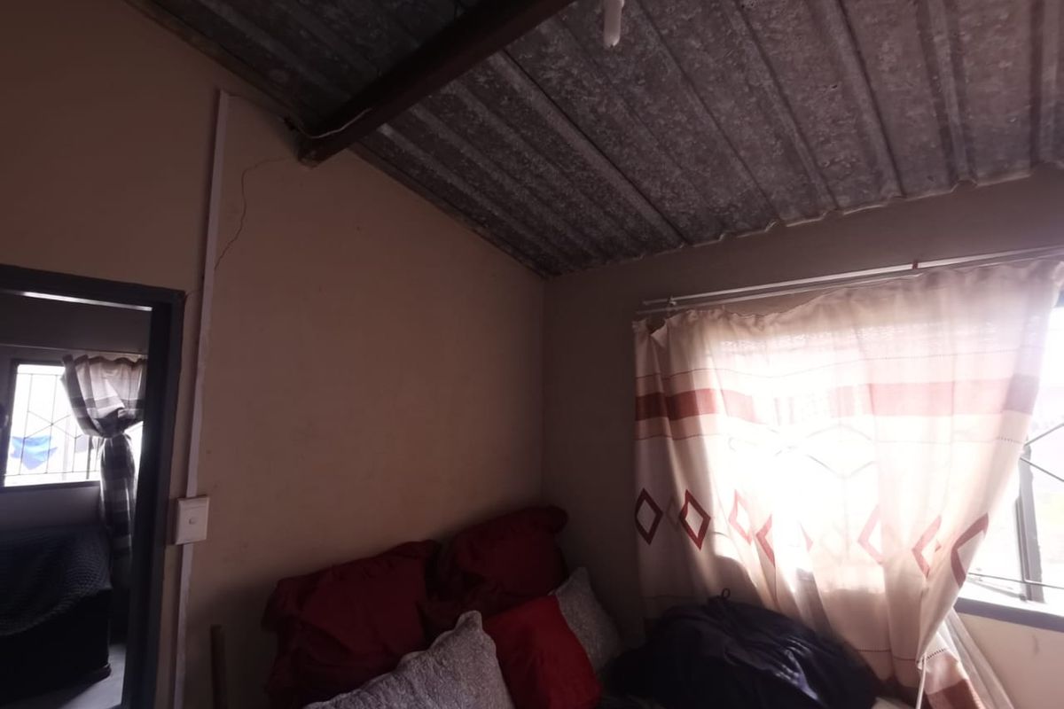 A bedroom featuring a corrugated metal ceiling and security bars on the windows, showing signs of wear including visible wall cracks.