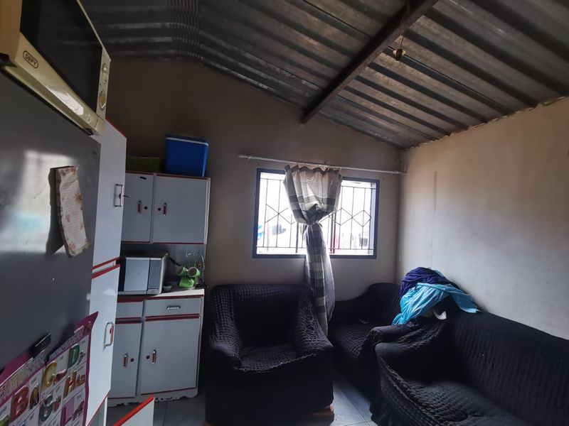 Open plan living and kitchen area featuring exposed corrugated metal roofing, tiled floors, and basic built-in cabinetry with security bars on the window.