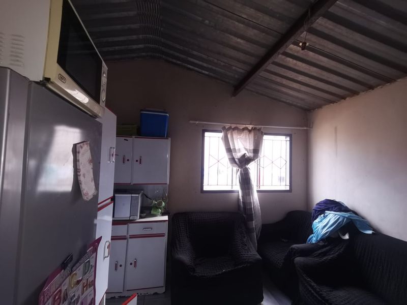 An open-plan living and kitchen area featuring basic built-in cabinetry, security bars on the window, and an exposed corrugated metal roof structure.