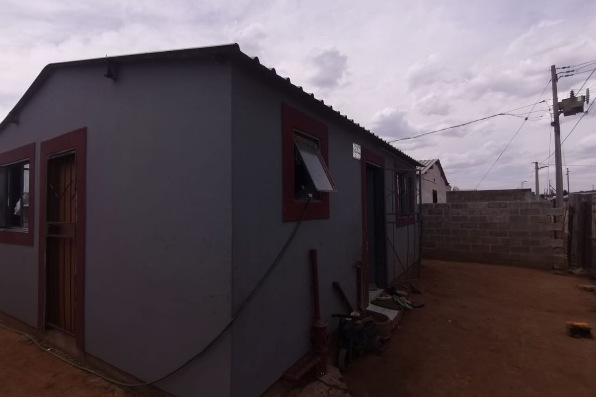 Rear view of a modest residential dwelling featuring a grey rendered facade, corrugated metal roofing, and a surrounding concrete block boundary wall.