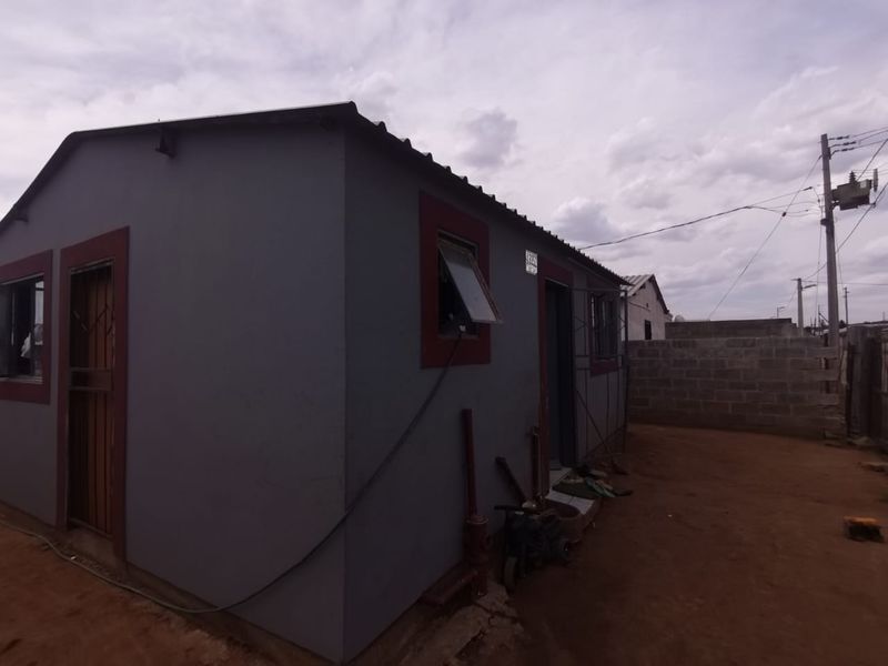 Rear view of a modest residential dwelling featuring a grey rendered facade, corrugated metal roofing, and a surrounding concrete block boundary wall.