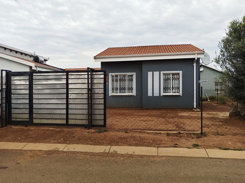 Front view of a single-story house featuring a tiled roof, security gates, and a metal driveway gate with an unpaved yard area.