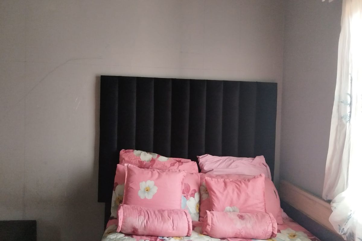 A bedroom featuring a dark upholstered headboard and a window with a curtain rail, showing signs of wear on the wall surfaces.