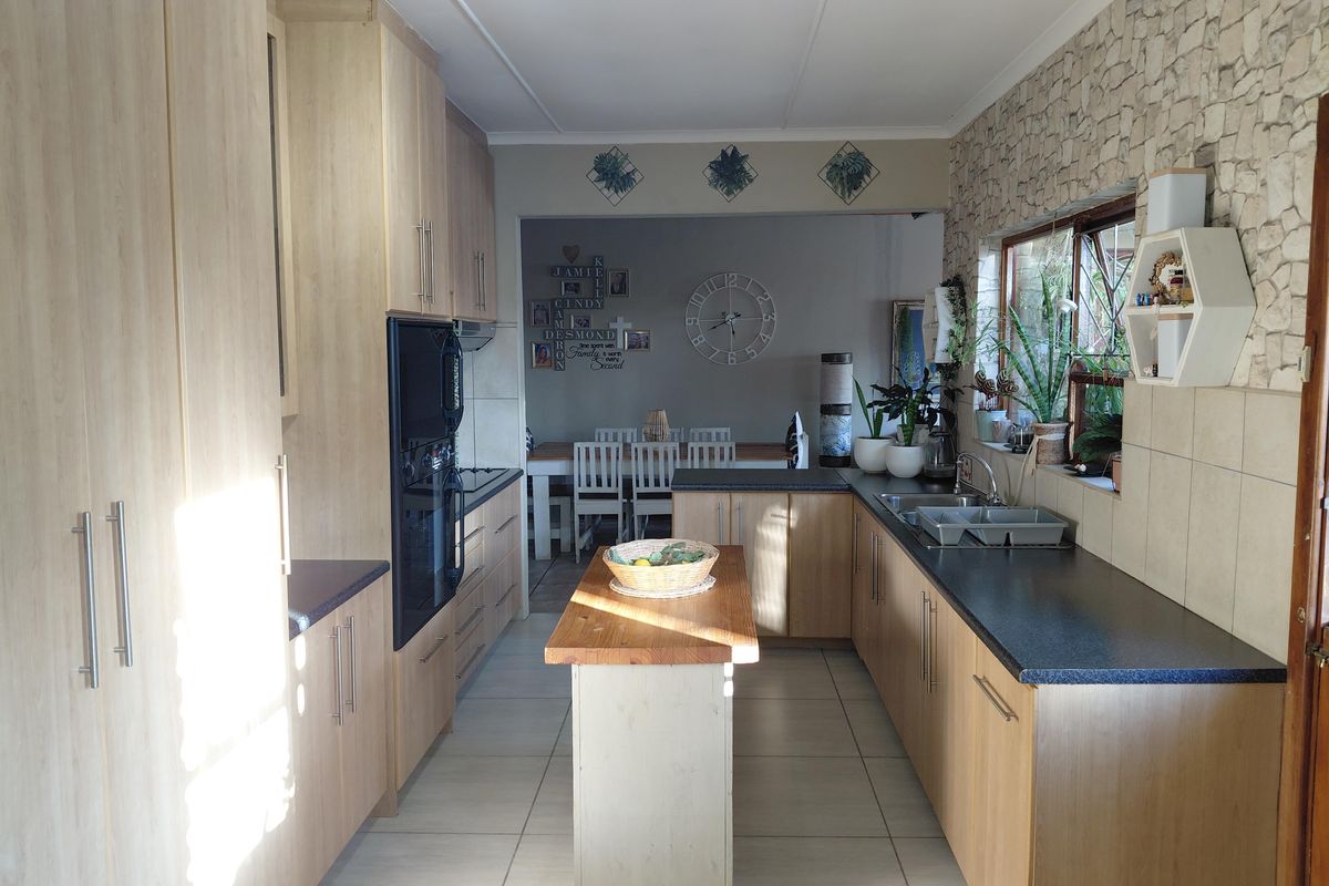 A functional kitchen featuring light wood cabinetry, dark laminate countertops, a central island, and tiled flooring, with an adjacent dining area visible.