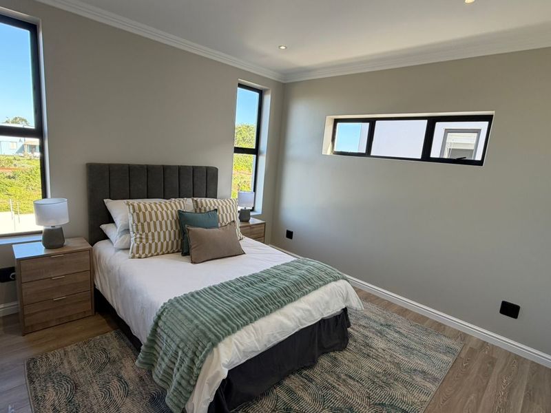 A modern, well-lit bedroom featuring neutral walls, laminate flooring, built-in cupboards, and large aluminium-framed windows providing ample natural light.