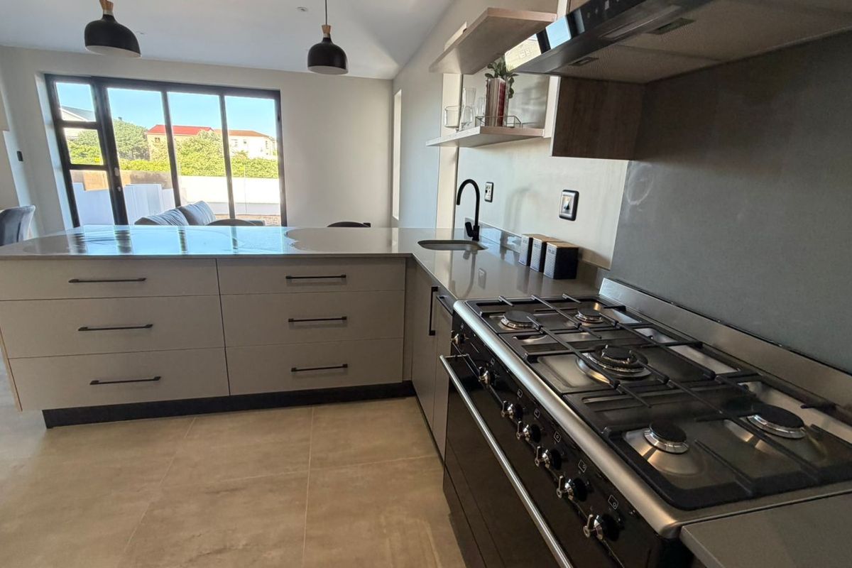 Modern kitchen featuring stone countertops, gas stove, extractor fan, and sleek cabinetry, seamlessly flowing into an open-plan living area with sliding glass doors.