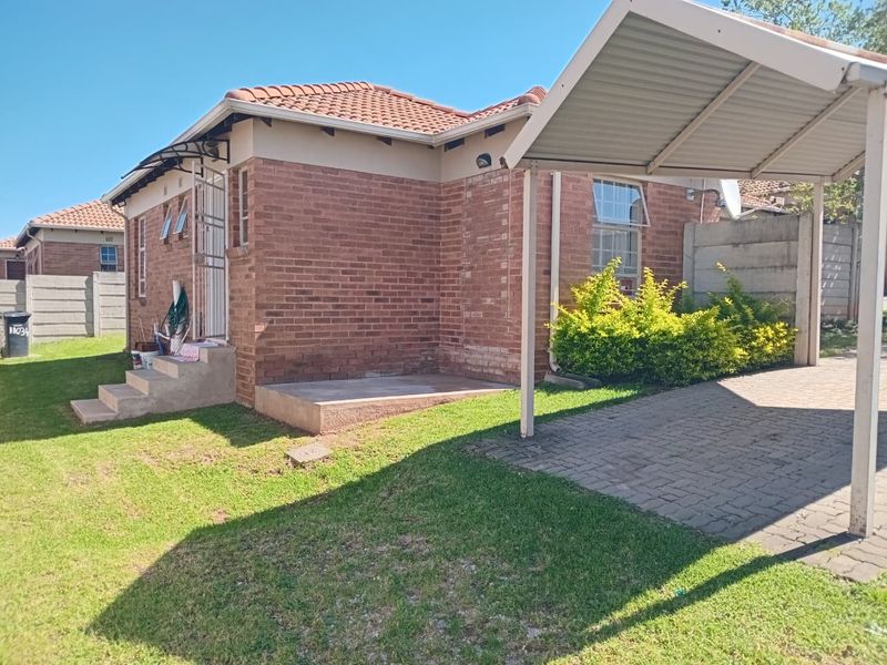 Exterior view of a facebrick townhouse featuring a tiled roof, a private carport, paved driveway, and a small surrounding garden area.