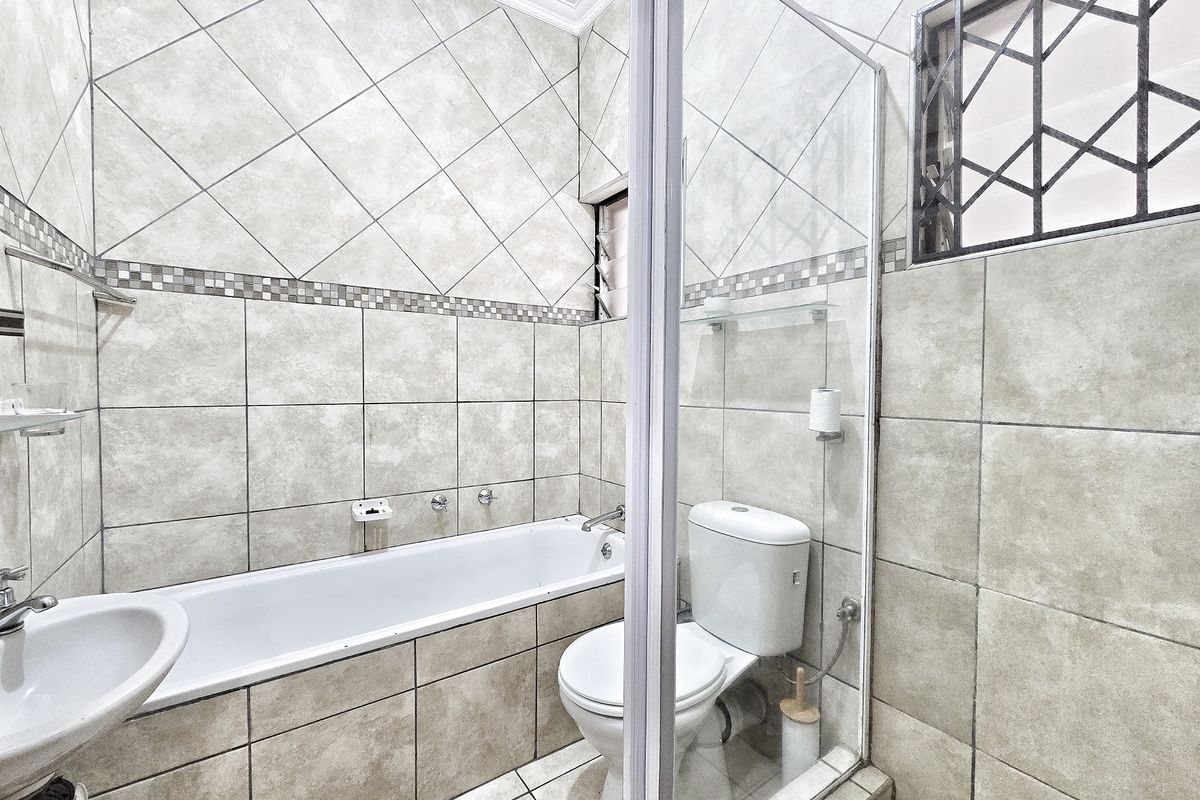 A functional bathroom featuring a bathtub, toilet, and glass shower screen with neutral tiled finishes and security bars on the window.