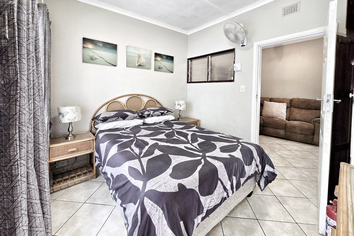 A bedroom featuring tiled flooring, a wall-mounted fan, and a louvered window, with an adjacent view into a living area.