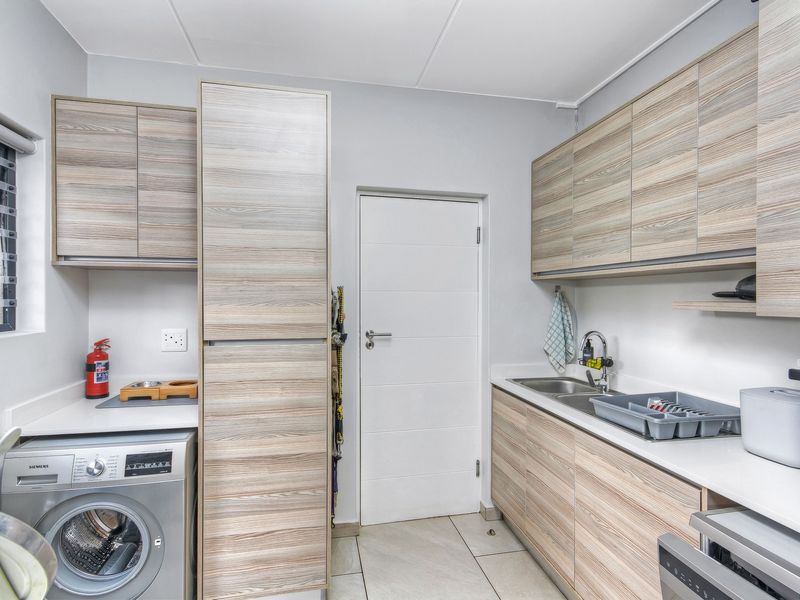A functional laundry room featuring light wood-grain cabinetry, stone-look countertops, a stainless steel sink, and space for a washing machine.