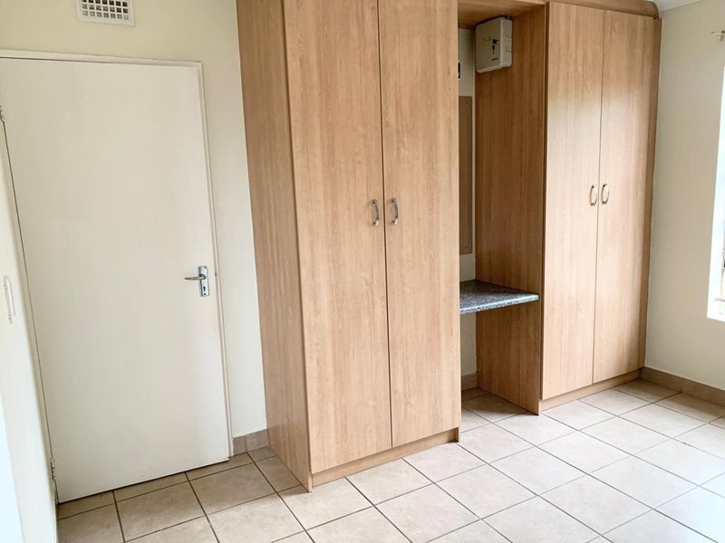 A clean, empty bedroom featuring light wood-finish built-in cupboards with an integrated desk area and neutral tiled flooring.