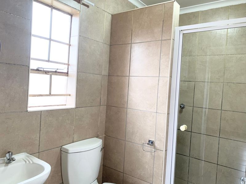 A functional bathroom featuring floor-to-ceiling ceramic tiling, a shower enclosure, a standard toilet, and a basin with a window providing natural light.