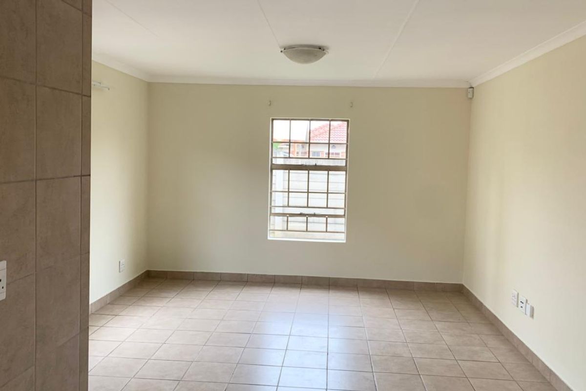 A bright, empty living room featuring neutral tiled flooring, a central ceiling light, and a window with security bars.