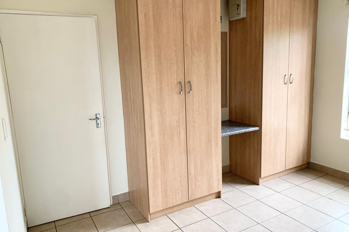A clean, empty bedroom featuring light wood-finish built-in cupboards with an integrated desk area and neutral tiled flooring.