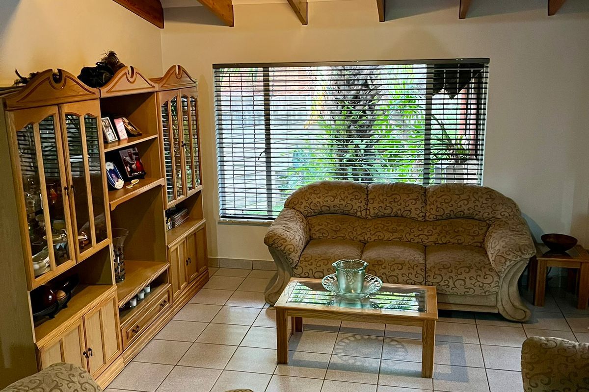 A comfortable living room featuring exposed timber ceiling beams, ceramic floor tiling, and large windows fitted with venetian blinds.
