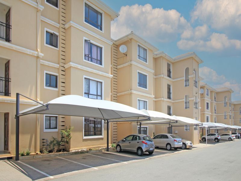 Exterior view of a multi-story apartment complex featuring paved parking bays with shade cloth carports and private balconies.