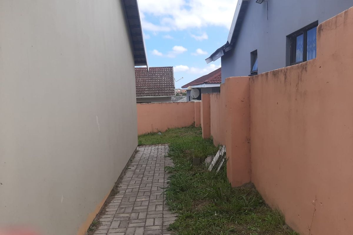 A narrow side passage featuring a paved walkway between the house exterior and a boundary wall, with some visible weathering and vegetation growth.