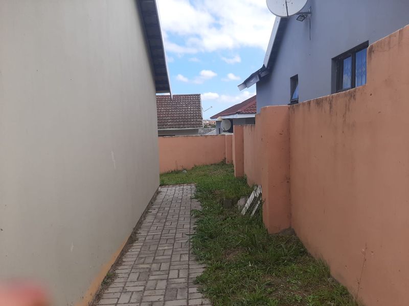A narrow side passage featuring a paved walkway between the house exterior and a boundary wall, with some visible weathering and vegetation growth.