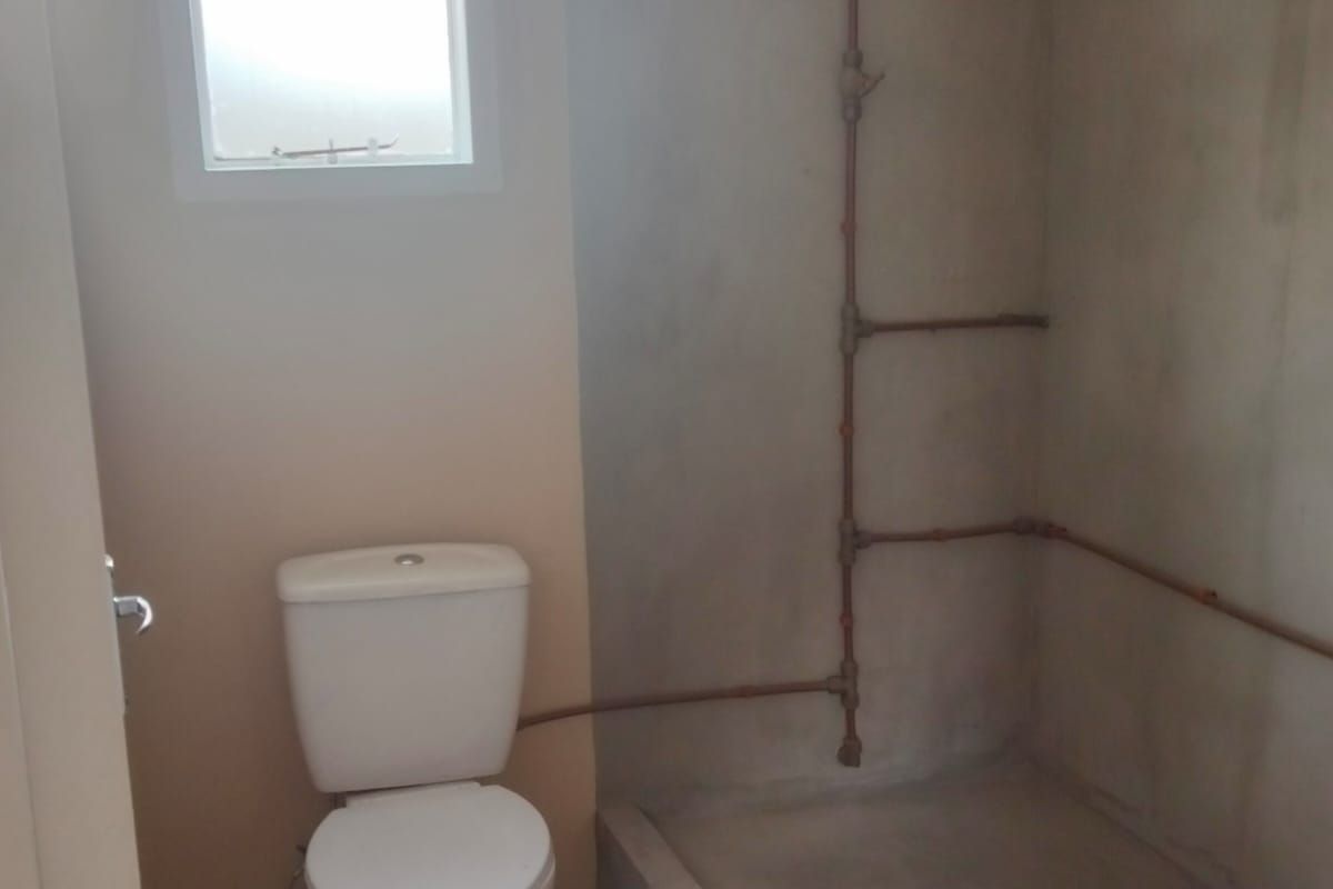 A basic bathroom featuring a toilet and a shower area with exposed copper piping and unfinished concrete walls.