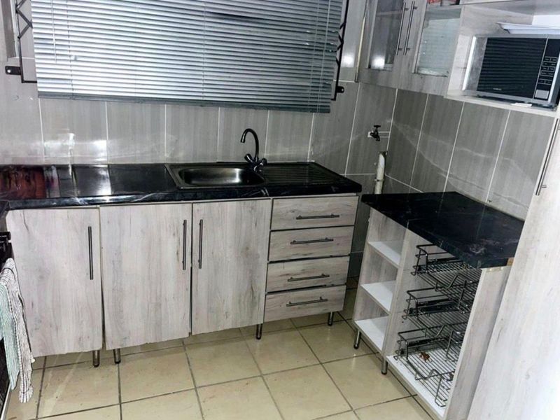 A functional kitchen featuring light wood-grain cabinetry, dark laminate countertops, a stainless steel sink, and tiled finishes throughout.