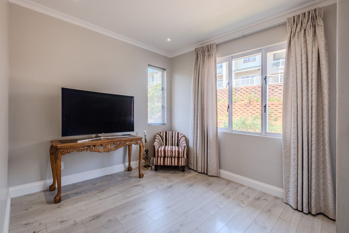 A bright living room featuring light-toned laminate flooring, neutral walls, cornicing, and large windows with integrated curtain tracks.