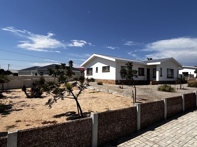 A single-story residential property featuring a paved driveway, white exterior walls with brick detailing, and a metal roof, set behind a precast boundary wall.