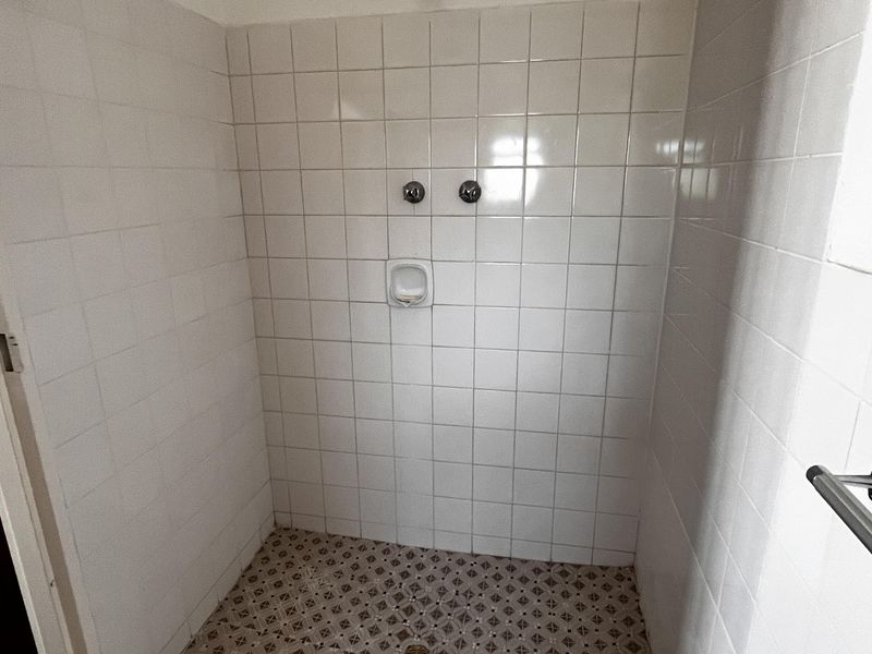A functional shower enclosure featuring white wall tiles, a patterned tiled floor, and basic chrome shower fixtures.